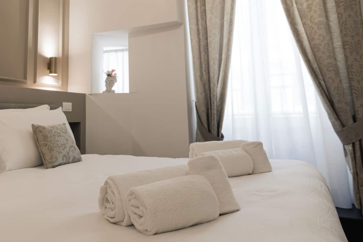 A neatly made bed with rolled towels and pillows, a bedside table with a vase, and a window with curtains.