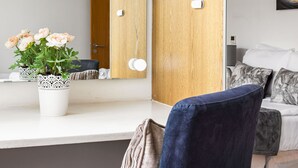Ein modernes Zimmer mit Schminktisch, einem beleuchteten Spiegel, einer Topfpflanze und einer Couch mit Kissen.