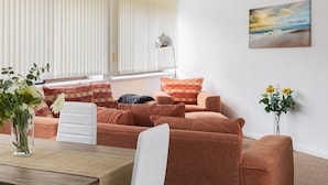 Ein modernes Wohnzimmer mit einer Sofagarnitur, einem Esstisch und einem großen Fenster mit Jalousien.