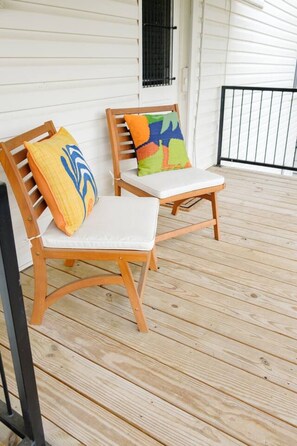 Two wooden chairs with cushions on a wooden deck, a white wall, and a window with a black shutter.