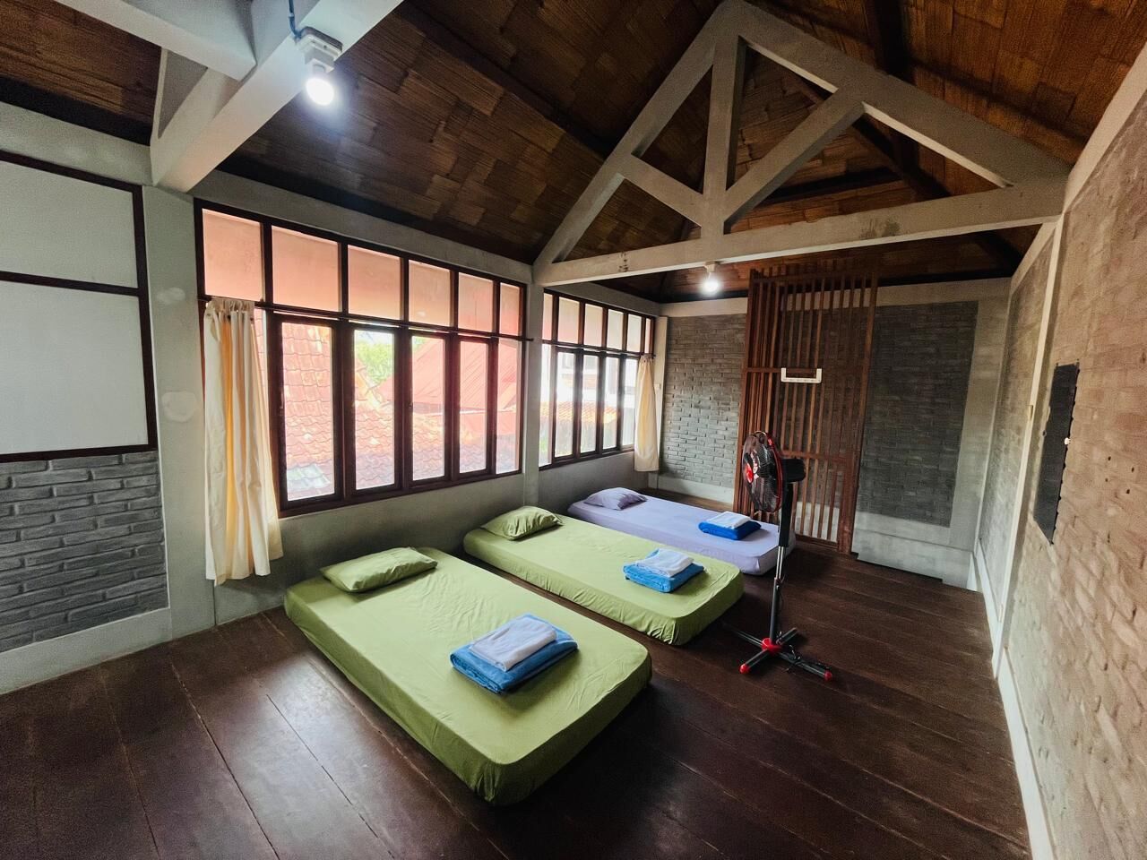 A room with two single beds, a wooden ceiling, brick walls, and large windows.