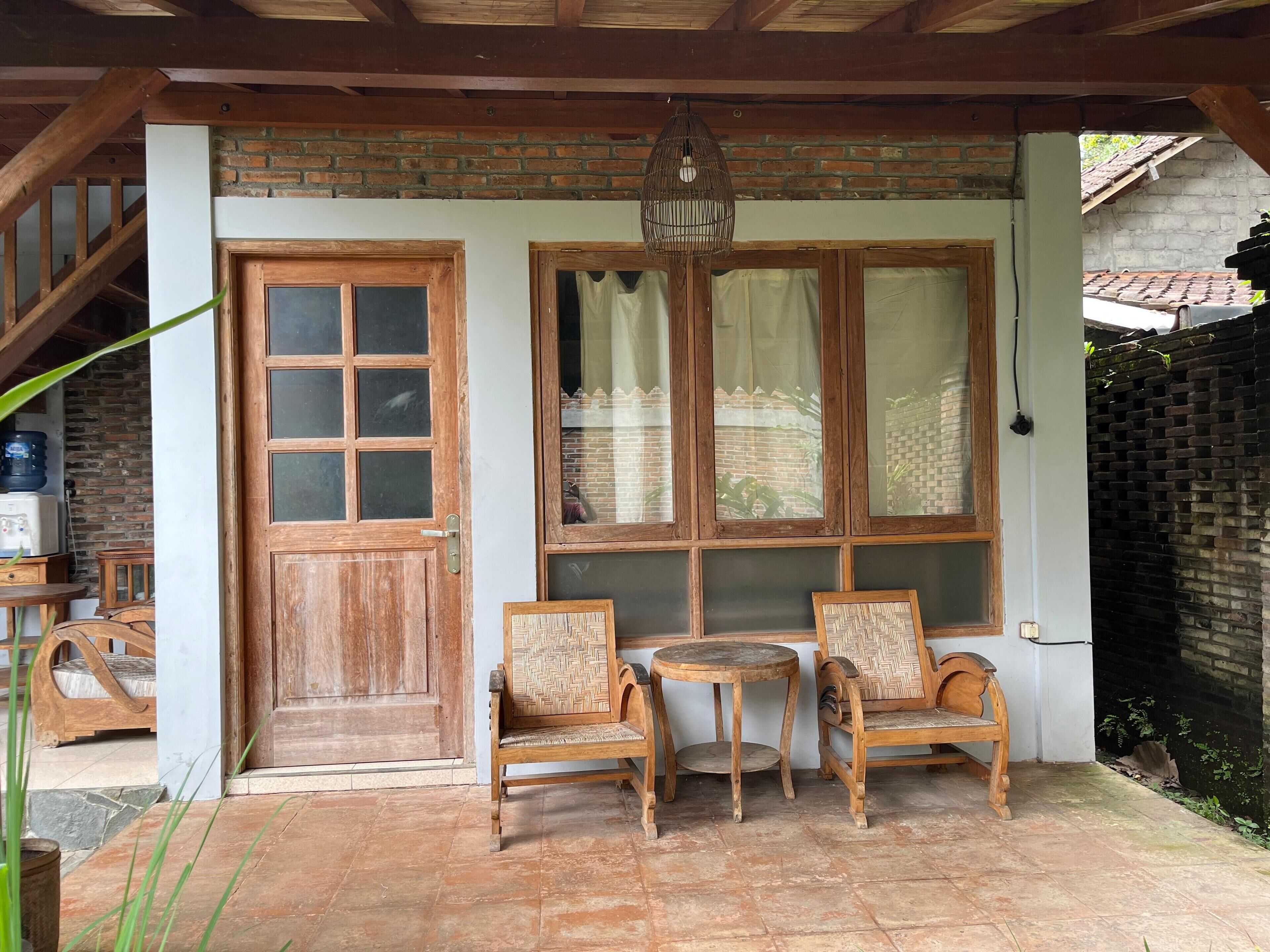 A covered patio with a wooden door, two wooden chairs, a small round table, and a hanging light fixture.