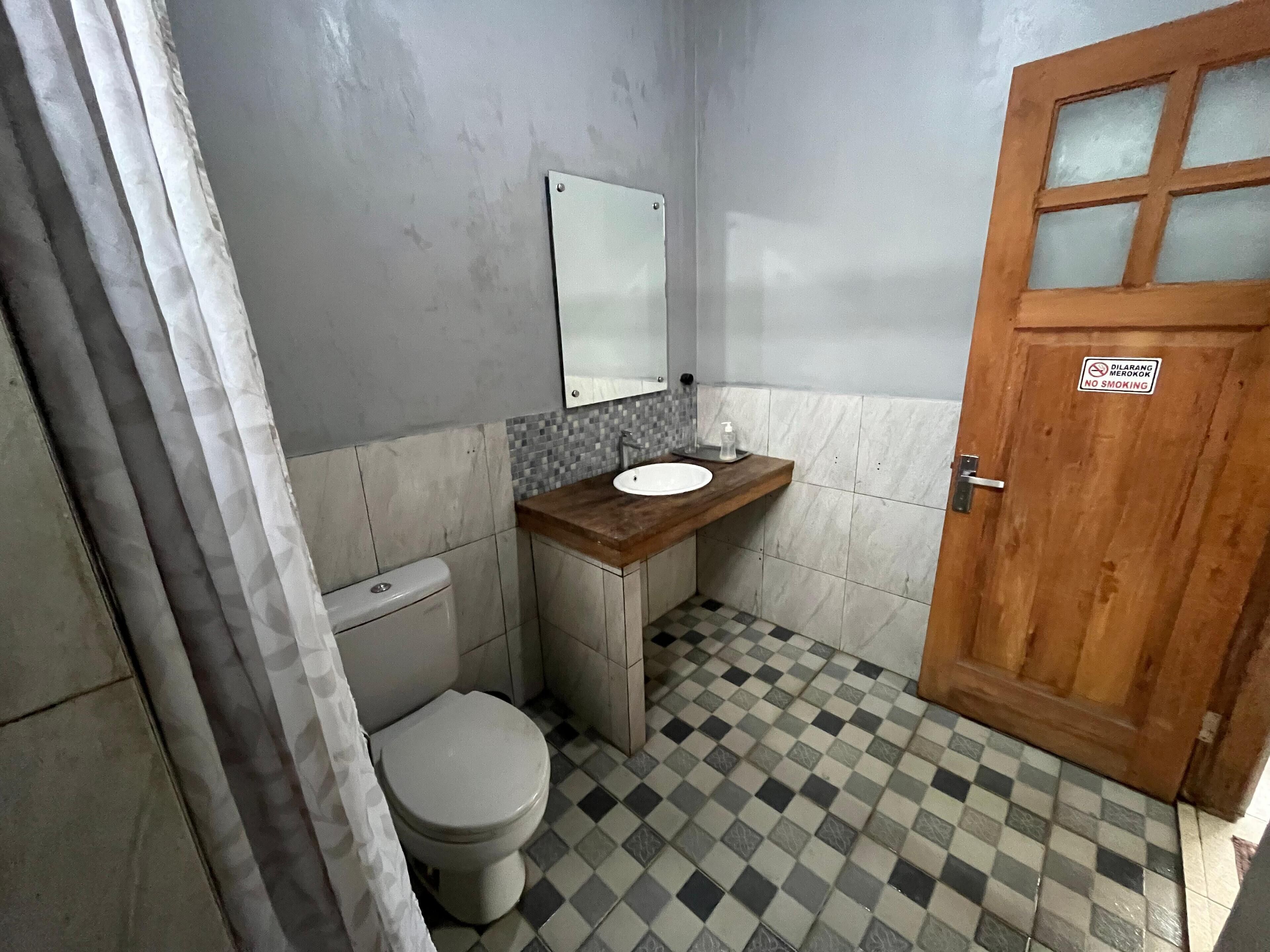 Een compacte badkamer met toilet, wastafel en spiegel. De vloer is zwart-wit geruit en de houten deur heeft een ruit.