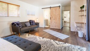 A modern living room with a grey sofa, a blue ottoman, and a wooden floor. There is a sliding door leading to another room. - Heated pool, hot tub, arcade, walk to town (Los Alamos)