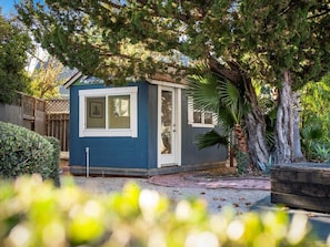 A small, single-story house with a blue exterior and white trim, surrounded by a garden with various plants and a large tree.