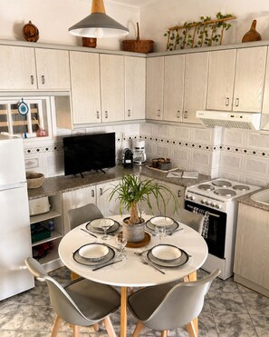 A kitchen with white cabinets, a round dining table with chairs, a stove, a TV, and a potted plant. - Stelios' Beachfront Retreat  (Paroikia)