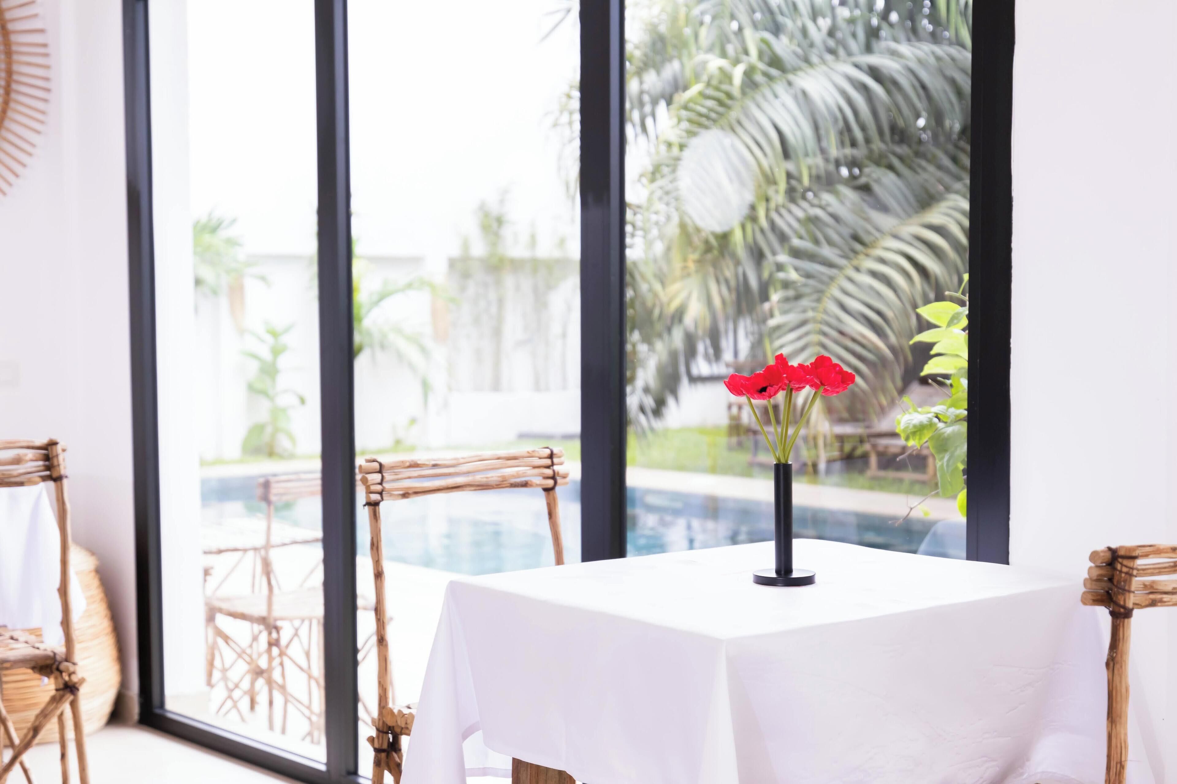 A dining area with a white table and chairs, a vase with red flowers, and a view of a swimming pool and palm trees through large windows.