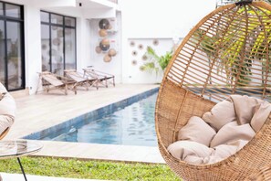 Zona de piscina con una silla de mimbre y una mesa de vidrio. Se observan sillas de descanso y un edificio moderno con amplias ventanas al fondo.