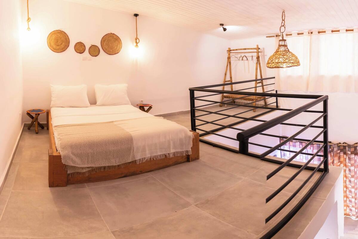 A bedroom with a bed, a small bedside table, a hanging light fixture, and a balcony railing.
