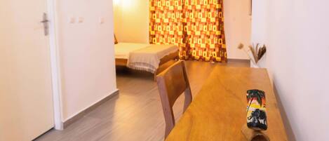 A room with a wooden dining table, a bed with patterned curtains, and a small wall-mounted light fixture.
