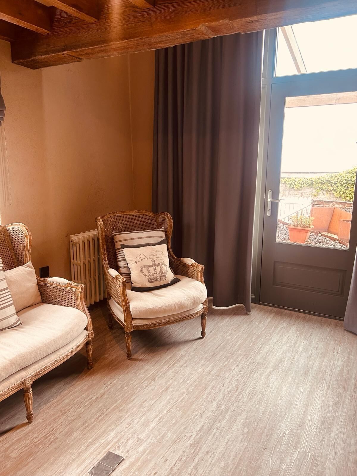 Een kamer met een houten plafond, een groot raam met gordijnen, een deur en twee rieten stoelen met kussens.