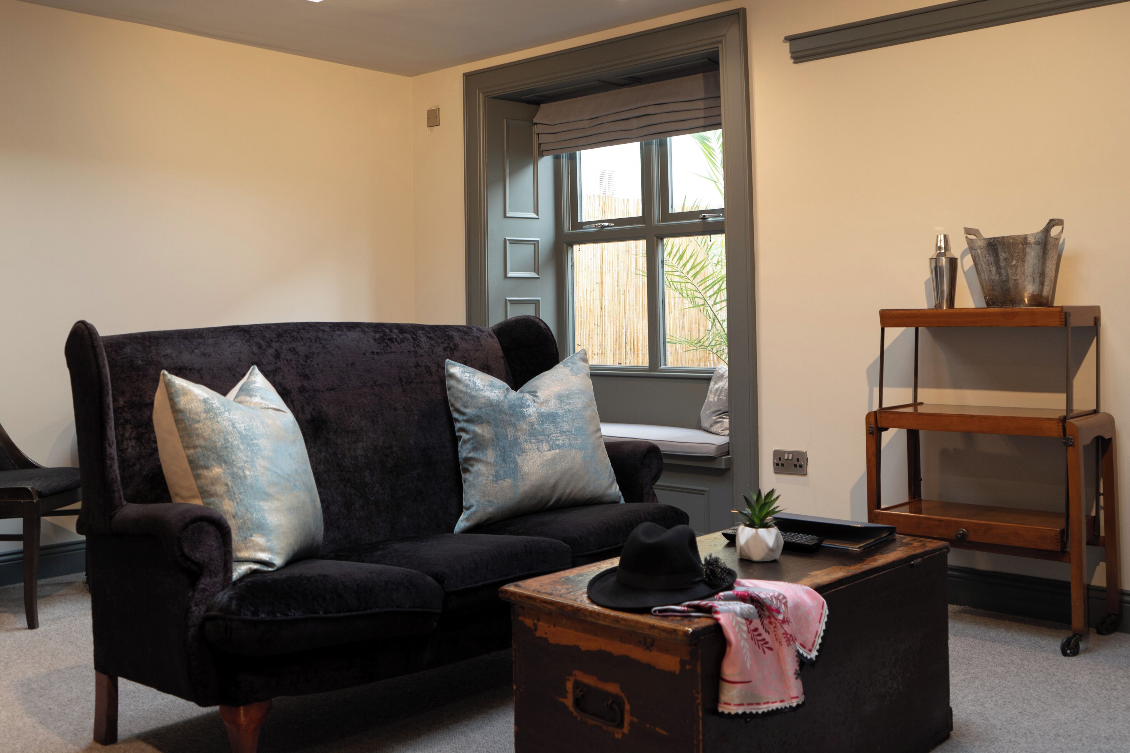 A living room with a black sofa, two blue pillows, a small table with a hat, a plant, and a wooden shelf with decorative items.