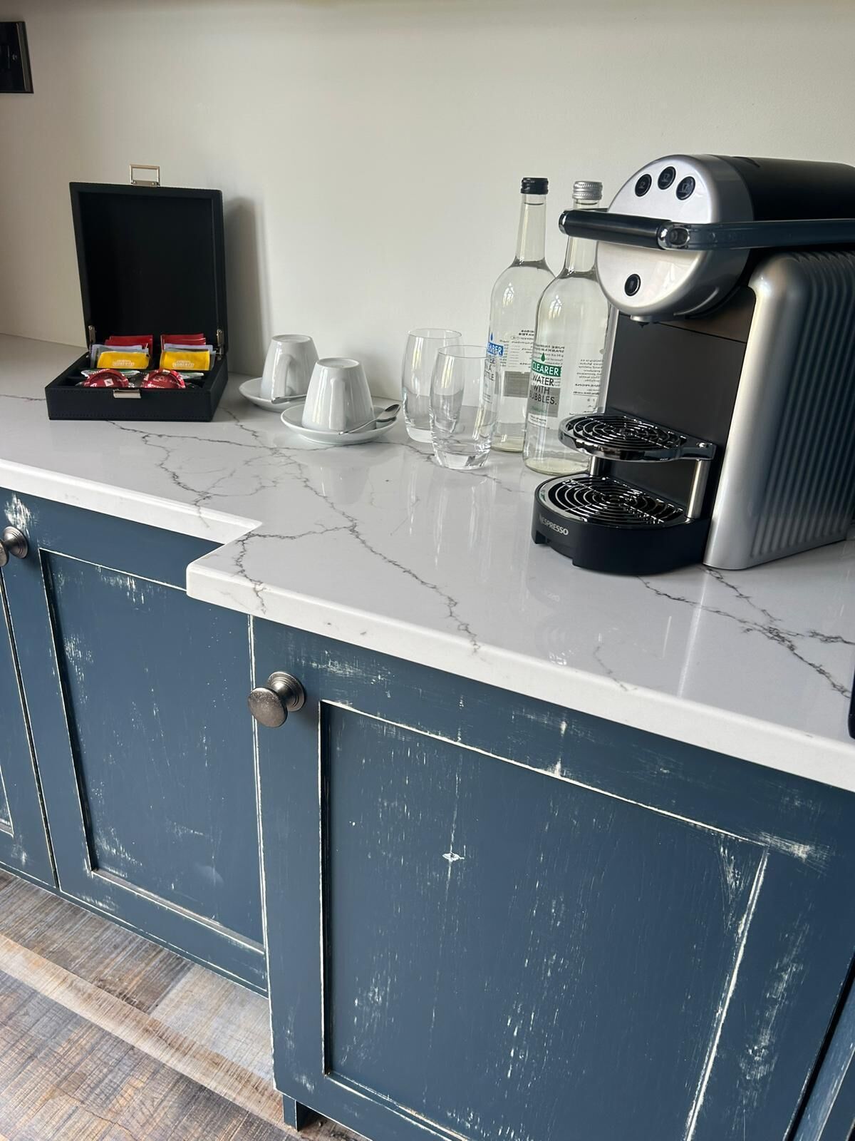 Una encimera de cocina con una cafetera, una caja de cápsulas de café y varios vasos.