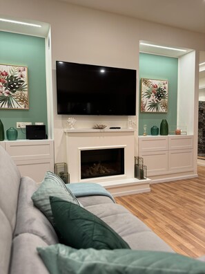 Ein modernes Wohnzimmer mit einem Flachbildfernseher über einem Kamin, einem grauen Sofa mit grünen Kissen und dekorativer Wandkunst.