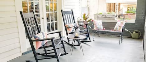 A covered porch with rocking chairs and a table, featuring a view of a house and garden.