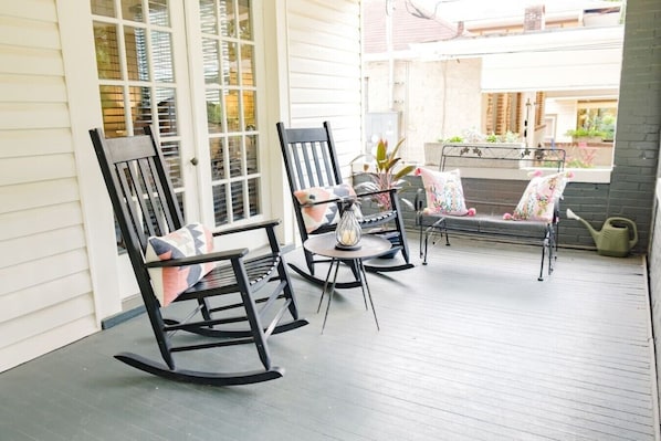 A covered porch with rocking chairs and a table, featuring a view of a house and garden.