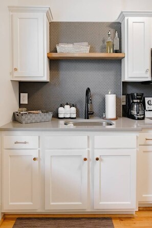 Una cocina con armarios blancos, salpicadero de azulejos grises, grifo negro y cafetera.