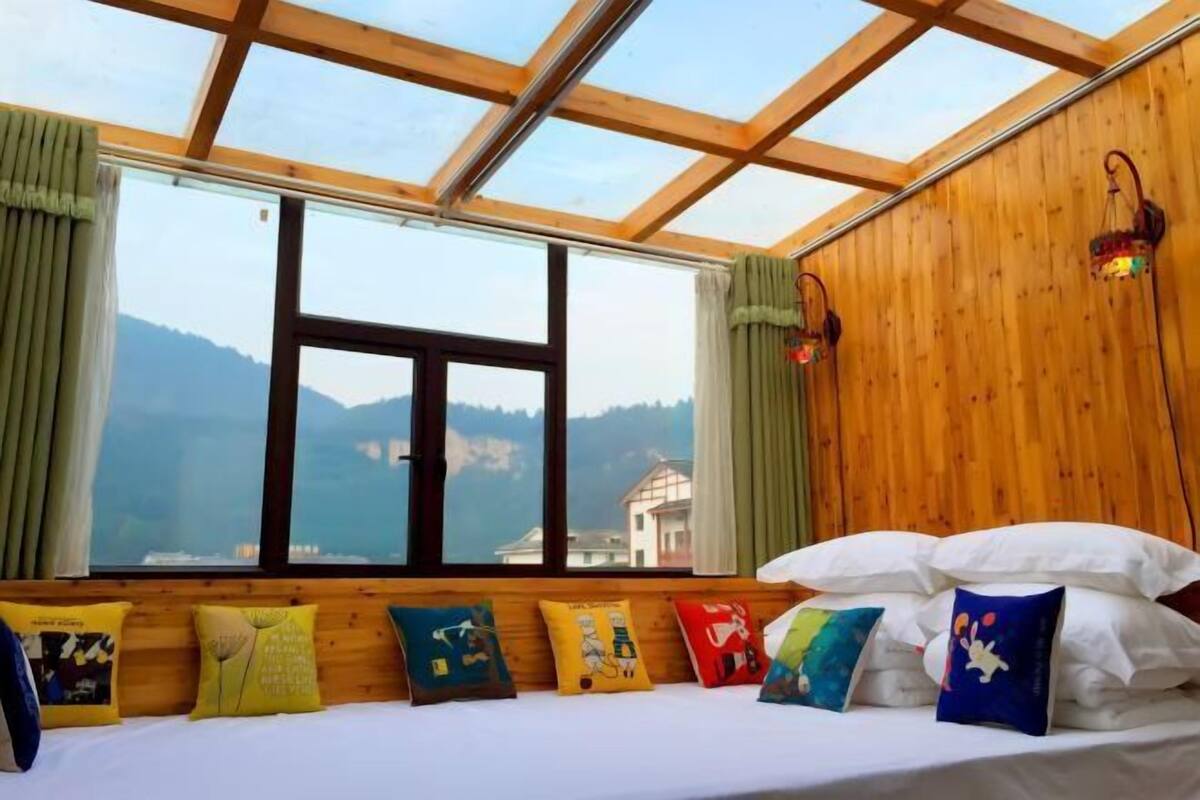 A room with a bed, colorful pillows, a wooden ceiling, and a view of mountains through large windows.