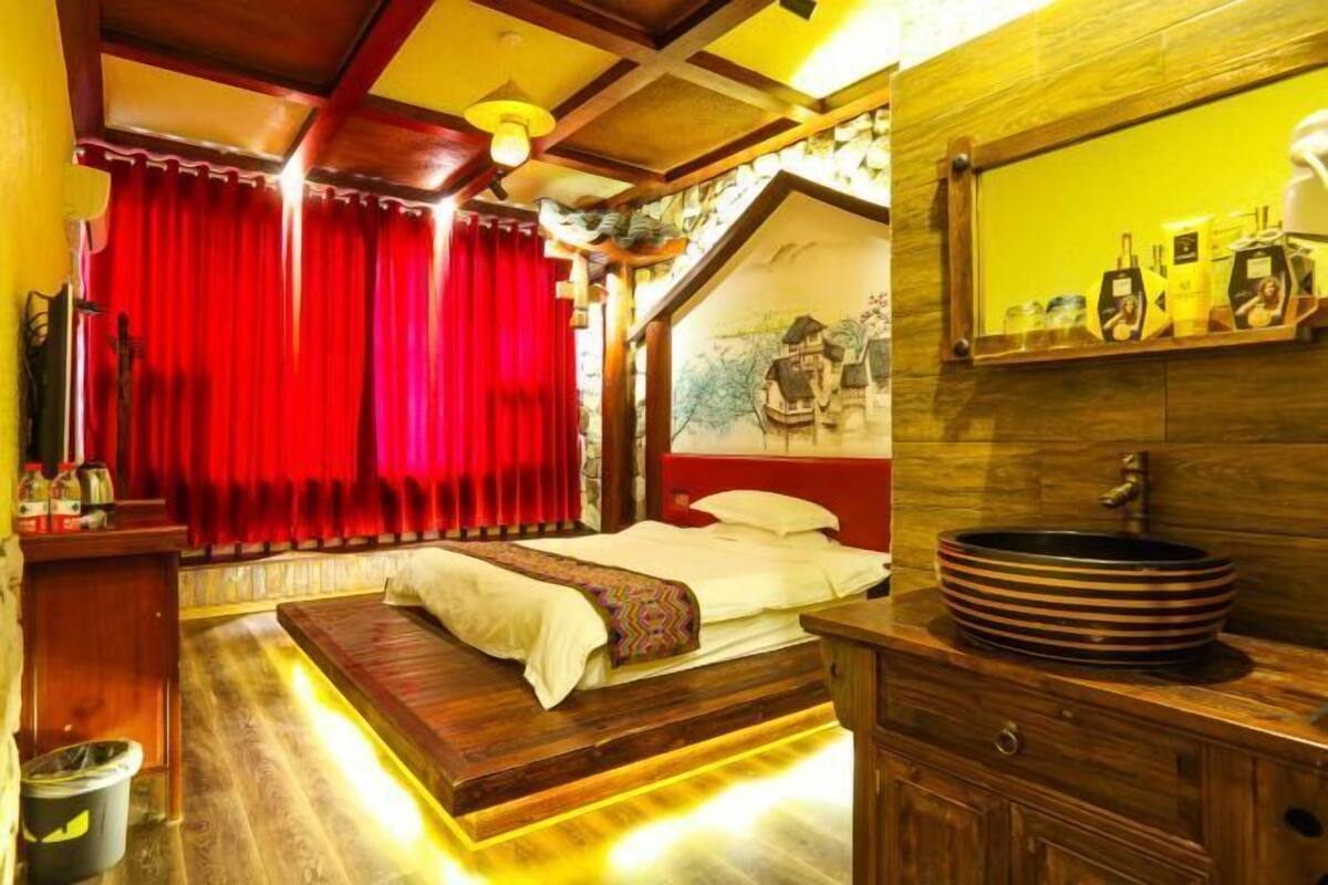 A bedroom with a bed, red curtains, a wooden headboard, a large mirror, and a wooden vanity with a sink.