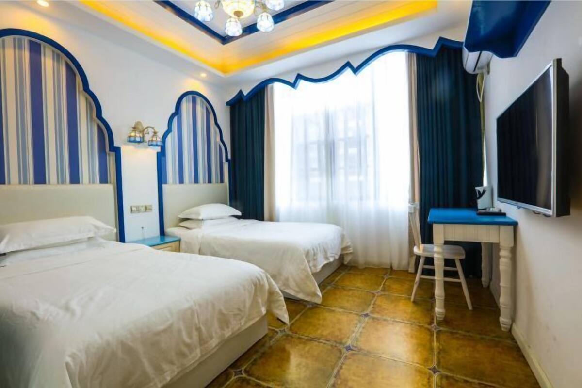 A hotel room with two beds, a desk, a TV, and a window with curtains.