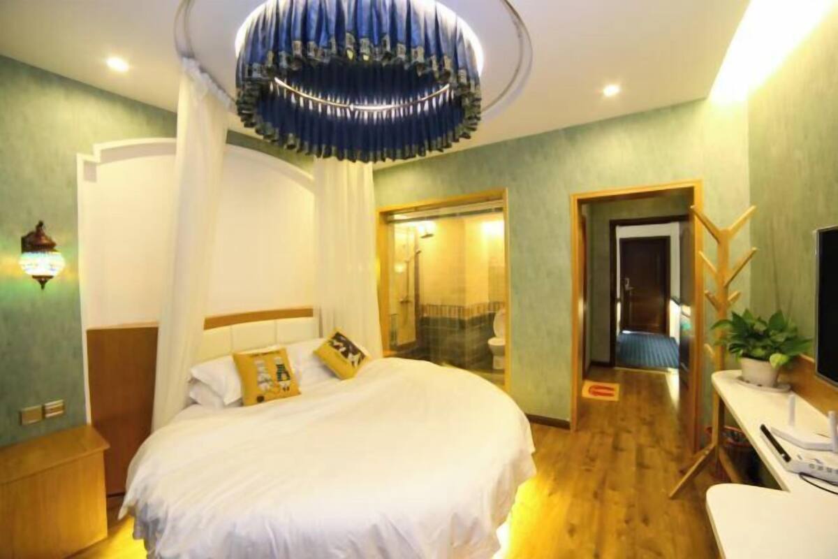 A hotel room with a large bed, a desk, a chair, a television, and a bathroom visible through a glass door.