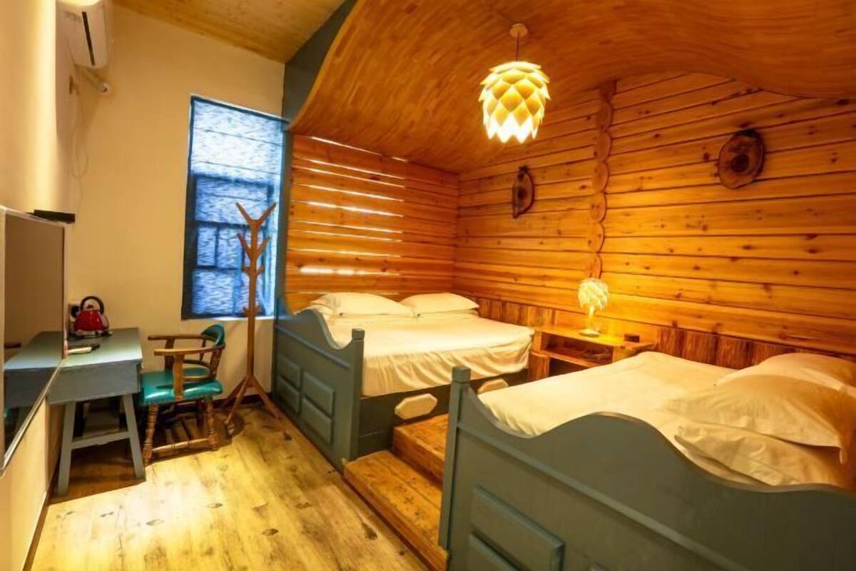 A wooden cabin room with bunk beds, a desk, a chair, a window, and a hanging light fixture.