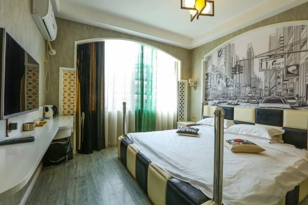 A hotel room with a bed, a TV, a window with curtains, and a cityscape mural on the wall.