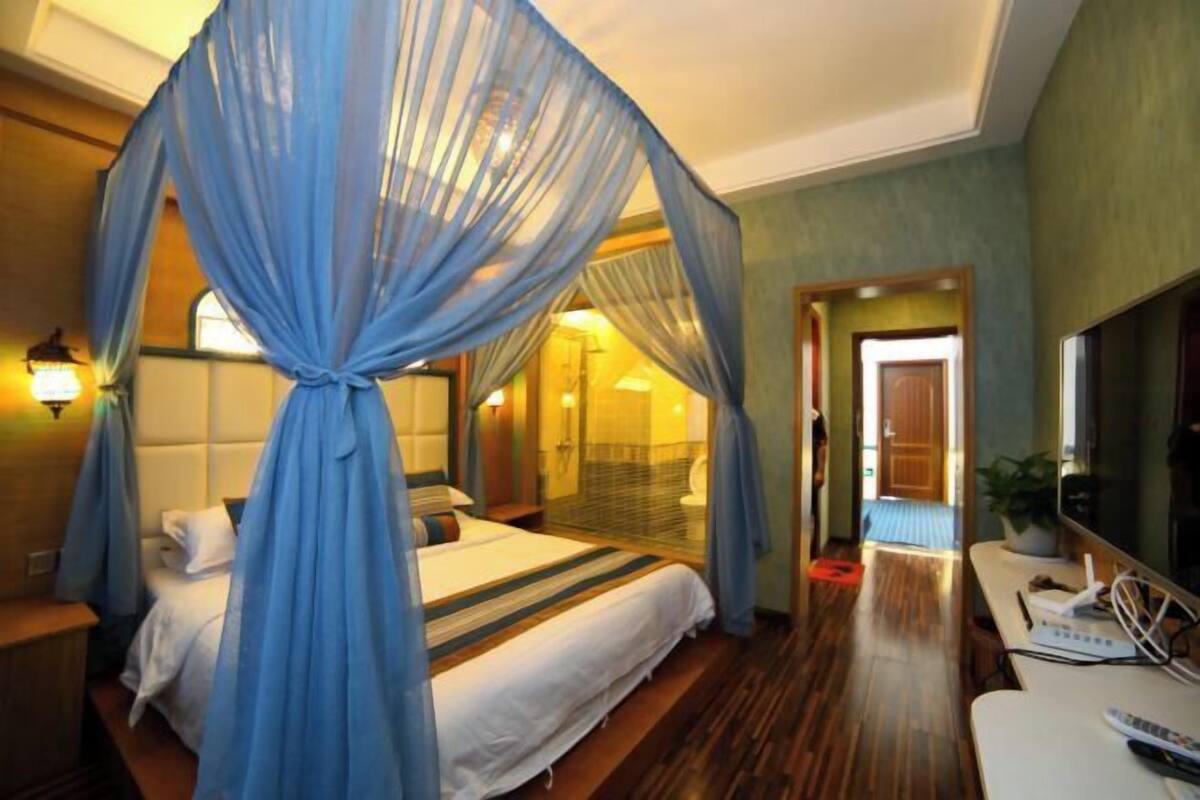 A hotel room with a bed, a desk, a chair, a TV, and a bathroom visible through a glass door.