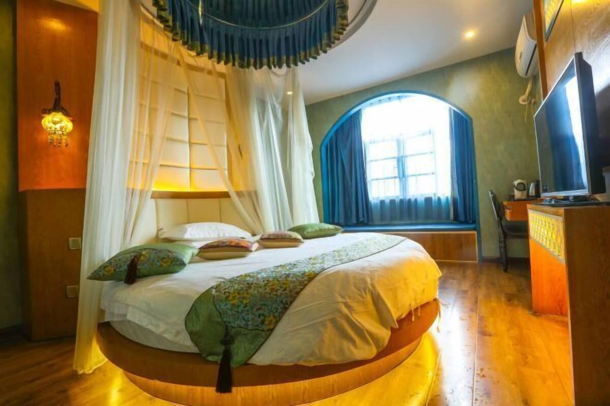 A circular bed with a canopy, a bedside table, a lamp, and a window with curtains.