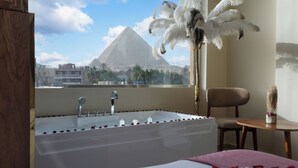 A bedroom with a bathtub, a bed with rose petals, a chair, and a view of a pyramid.