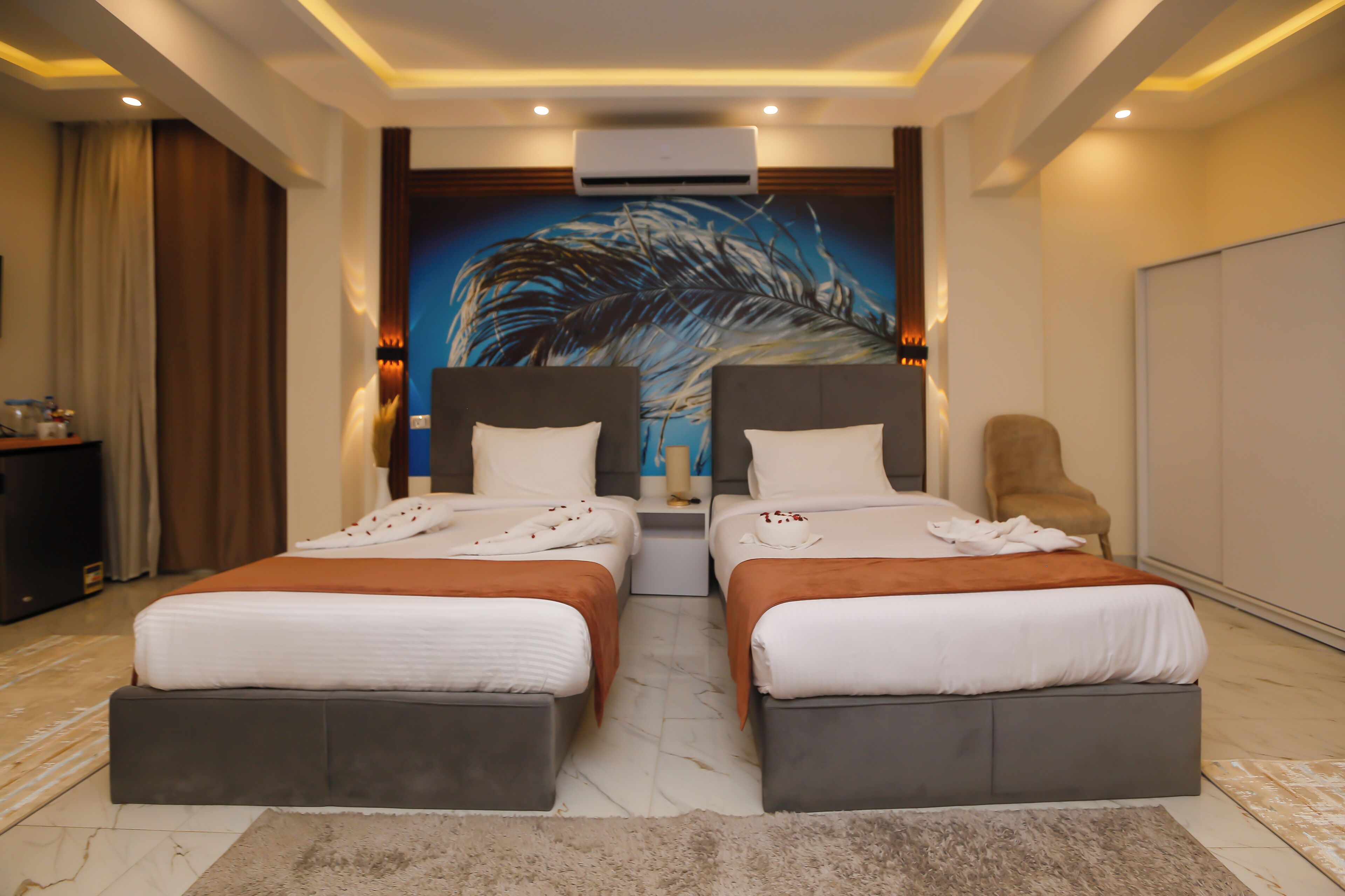 A hotel room with two beds, a large wall mural, an air conditioning unit, and a seating area with a chair.