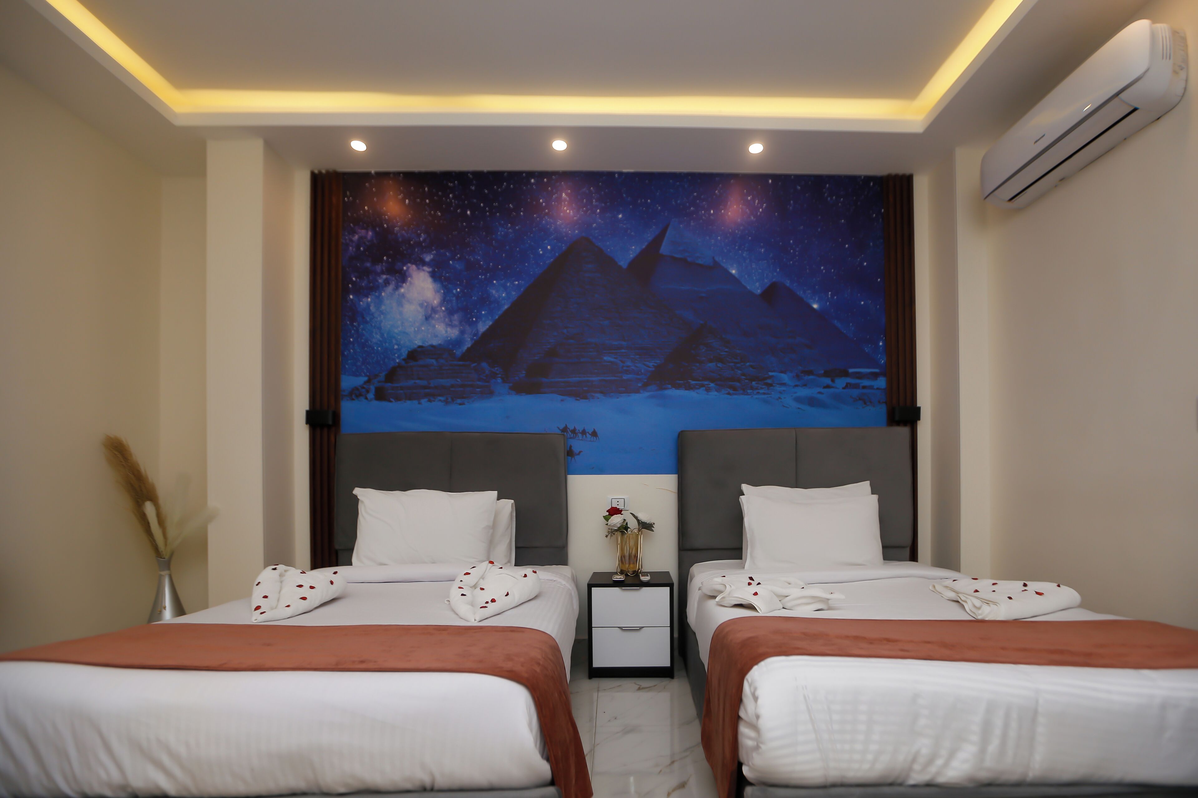 A hotel room with two beds, a nightstand, a wall tapestry of pyramids, and an air conditioning unit.