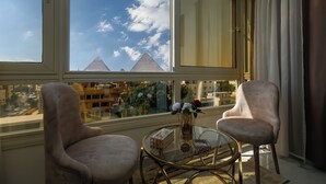 A room with a view of pyramids, two armchairs, a glass table with decorative items, and a window with curtains.