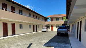 A courtyard with a paved walkway, a parked car, and a building with multiple doors and windows. - Apartamento Essecial Simplu (Maracanau)