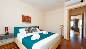 A neatly made bed with a teal bedspread and white linens, a framed artwork above the bed, and a modern air conditioning unit on the wall.