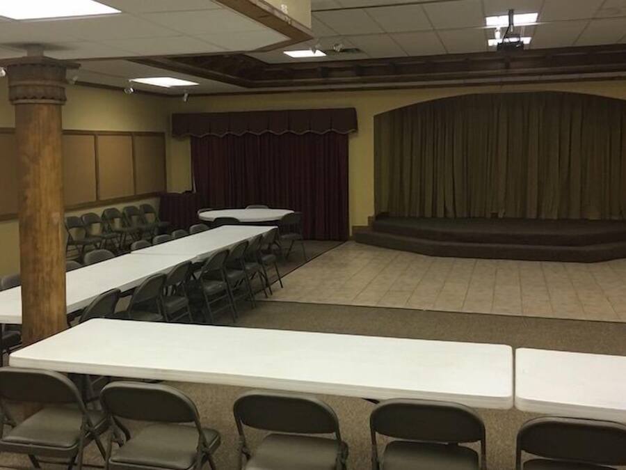Uma sala de conferências com mesas compridas e cadeiras, um palco com cortinas e um pilar de madeira.