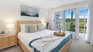 Ein geräumiges Schlafzimmer mit einem großen Bett, einem Nachttisch mit Lampe und Blick auf einen Garten durch eine Schiebetür.