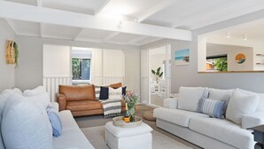 Ein modernes Wohnzimmer mit einer braunen Ledersofa, einem weißen Sofa und einem weißen Sessel. Auf einem Korbtisch stehen eine Flasche und Gläser. Im Hintergrund ist ein Fenster mit Blick auf Grünpflanzen zu sehen.