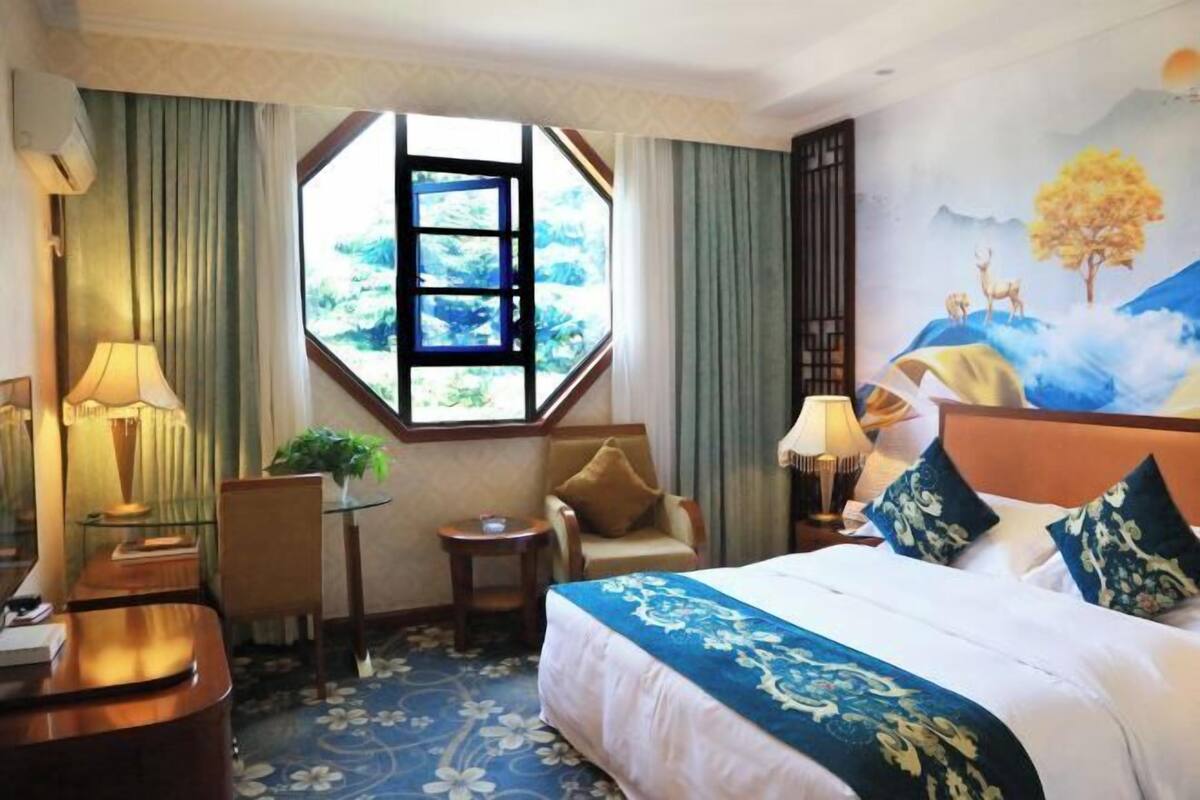 A hotel room with a large bed, a desk, a chair, a small table, and a window with a view of greenery.