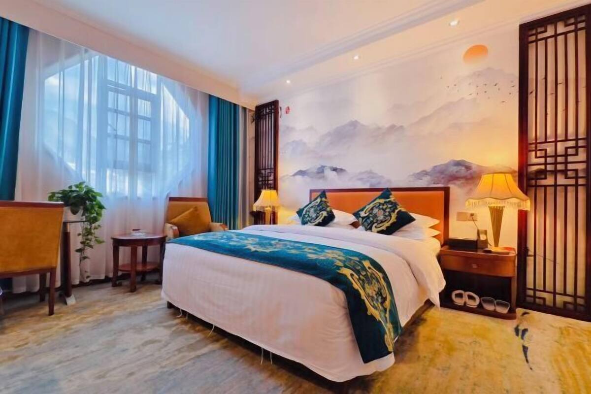 A hotel room with a bed, a desk, a chair, a small table, a lamp, and a wall mural.