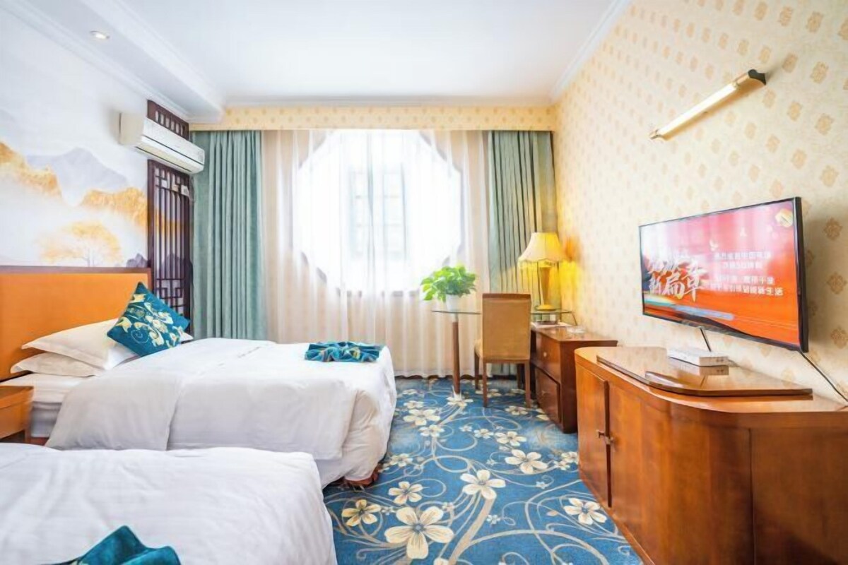 Una habitación de hotel con dos camas, un escritorio con silla, televisión y ventana con cortinas.