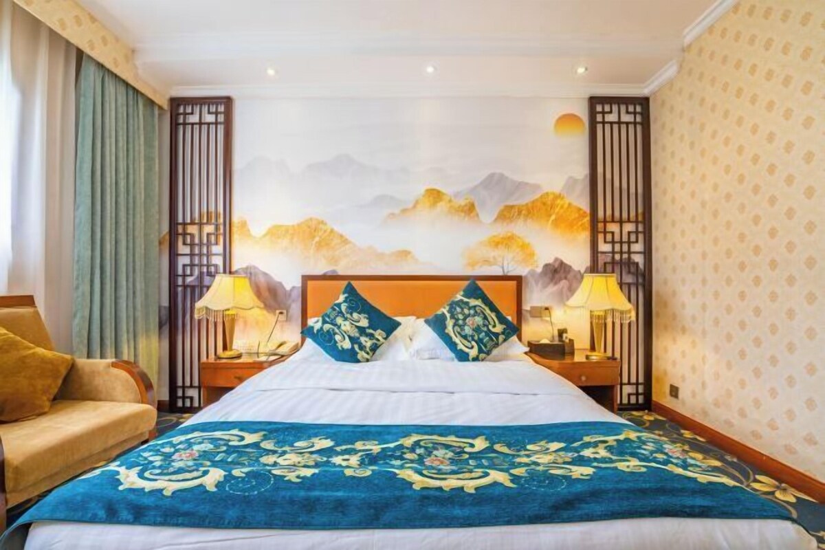 Habitación de hotel con una cama grande, dos mesitas de noche, un sofá y un mural en la pared que representa montañas y un atardecer.