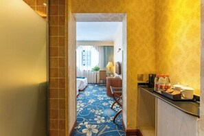 Un pasillo de hotel con alfombra floral, una cama, un escritorio con lámpara y un mostrador con productos para el desayuno.