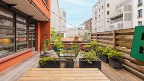 En smal bygård med træterrasse, plantekasser og en hængestol.