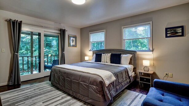 A bedroom with a large bed, two bedside lamps, a nightstand, a chair, and a view of the outdoors through a sliding door. - Creekside, spacious home for 8; 9 mi. Highlands, close Sky Valley, GA (Scaly Mountain)