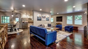 A spacious living area with a blue sofa, a wooden coffee table, and a dining table with chairs. There is a kitchen in the background with modern appliances. - Creekside, spacious home for 8; 9 mi. Highlands, close Sky Valley, GA (Scaly Mountain)
