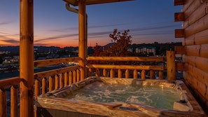 A wooden deck with a hot tub and a view of a cityscape at dusk.