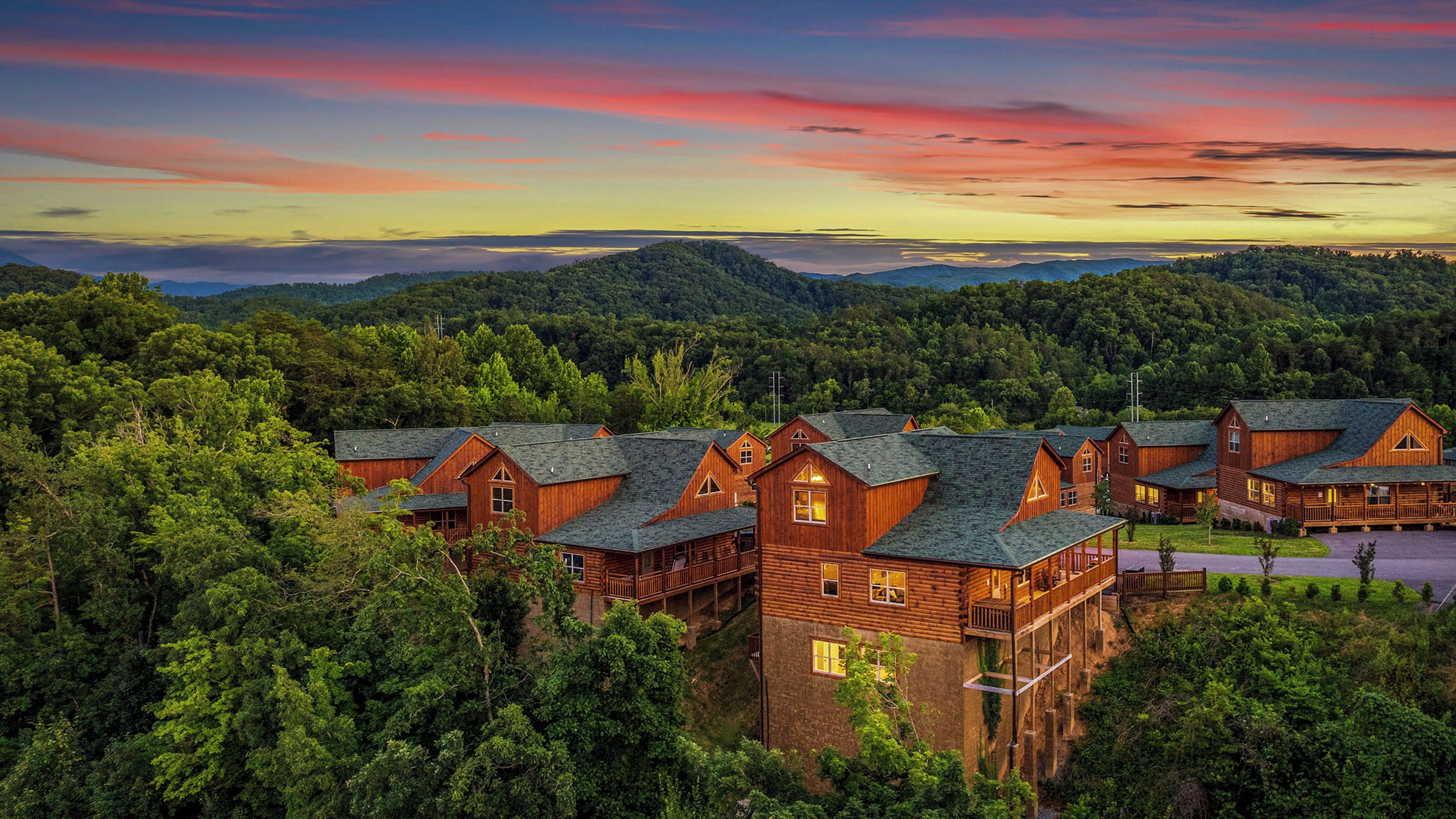 A lodge with multiple wooden cabins surrounded by dense forest, set against a backdrop of mountains and a colorful sky.