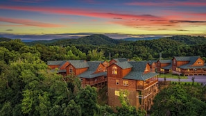 A lodge with multiple wooden cabins surrounded by dense forest, set against a backdrop of mountains and a colorful sky.