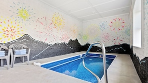 A swimming pool with a mountain mural on the wall, a chair with towels, and a ceiling with recessed lighting.